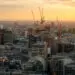 A city skyline at golden hour, with several orange construction cranes rising above a mix of modern glass office buildings and older stone architecture, hazy hills visible in the distance.